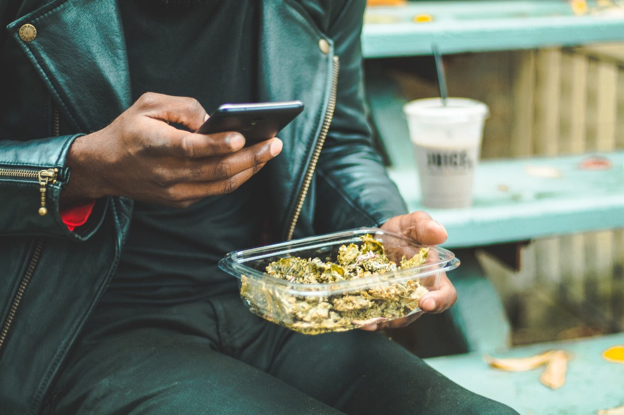 Hip man in black leather jacket holding container of Chicago Kale Chips in one hand while looking at his phone in his other.